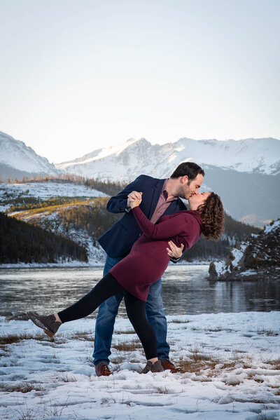 A couple kisses while dancing in the snow beside a lake, with snowy Colorado mountains in the background, captured beautifully by a Denver Colorado wedding photographer.