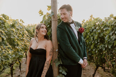 A bride in a black dress smiles up to her groom in a green jacket as they lean on a pole in a vineyard