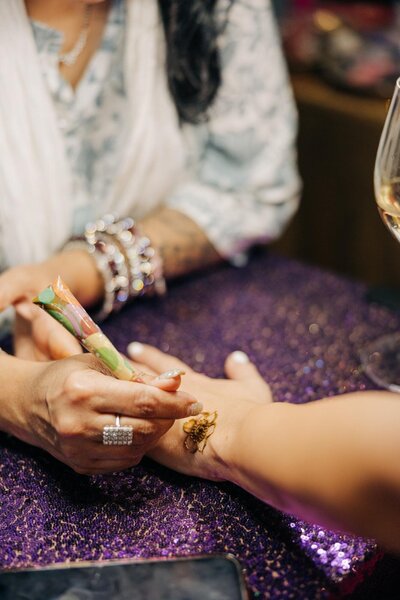Applying a henna design on a hand over a sequined purple surface.