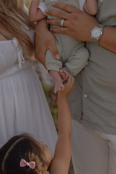 Family moment with a toddler reaching up to touch their newborn sibling’s foot while being held by the parents