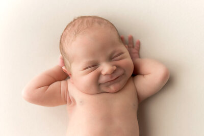 Newborn baby stretches an grimaces with hands behind head.