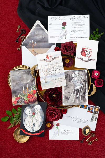 Elegant Halloween-themed wedding invitation suite by E Stokes Creations featuring detailed illustrations of skeletons, red roses, and gothic details, including candelabras and unique accents, perfect for a gothic romance-themed wedding.
