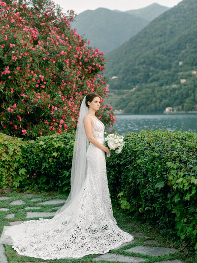 Bride in white lace bridal dress and veil holding white floral bouquet by Lake Como Wedding Italy