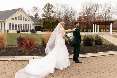 The bride approaches her groom from behind, her veil trailing behind her as she reaches out to tap his shoulder. The groom, dressed in a rich green suit, stands still, moments away from seeing his bride for the first time on their wedding day. The Pennsylvania venue's cream-colored siding and expansive windows provide a classic backdrop