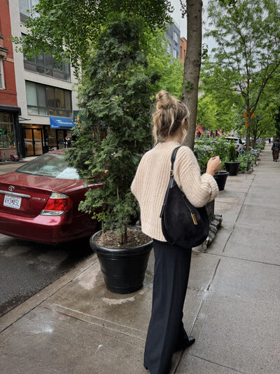 A woman wearing a beige knit sweater, black trousers, and a black suede tote bag walks down a tree-lined New York City street on a rainy day, showcasing a chic and casual urban outfit.
