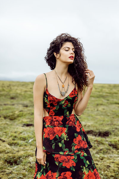 Editorial senior portrait of a curly-haired model in a bold black dress with red roses, standing in an open field on a cloudy day with wind softly blowing her hair.