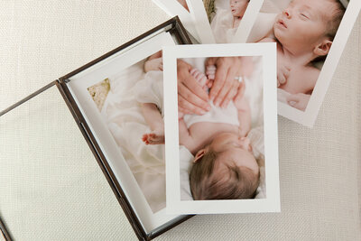 A glass print box with loose prints inside and to the right of the box. The loose prints show details of a newborn laying in a basket by Katie Stansfield Photography, a family photographer in Richmond.