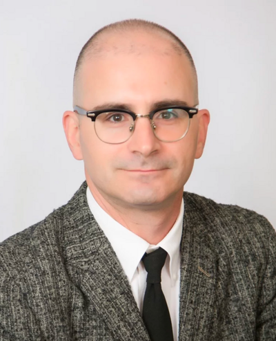 Headshot of Chris Cormier, a man with brown eyes, thin-rimmed glasses, a black and white tweed suit, white button down shirt and black tie