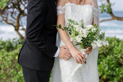 A close up of someone in a wedding dress holding a bouquet with their partner's hands around their waist 
