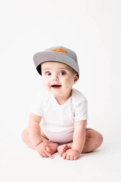 Baby in gray hat smiles for his photo on a white backdrop.