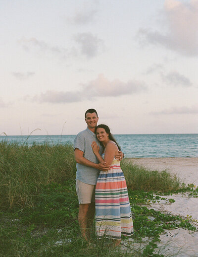 Stevie and husband embraces on a beach at sunset, surrounded by greenery. They smile warmly.
