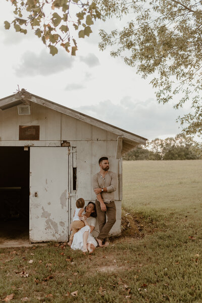 Outdoor family session of mama and dad leaning up against an old barn with their little toddler