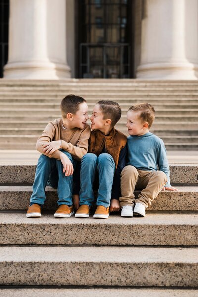 Three boys from a Pittsburgh family sit together on steps, one whispering into another's ear, captured by a talented photographer in Pittsburgh, PA.