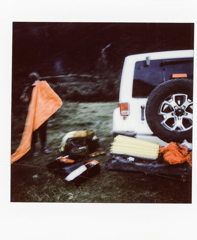 Polariod of JayneMayAgnes blowing up a camping mattress next to a white jeep in Alaska