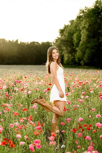 dark hair cuthbertson high school senior stands in red and pink poppy field wearing a white dress and brown boots at sunset