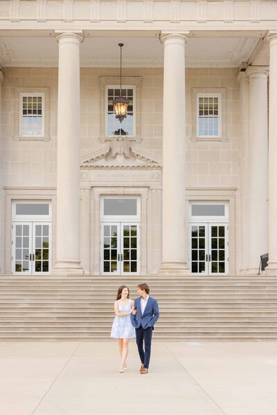 A couple walks arm in arm in front of the grand entrance of the Chicago History Museum, framed by tall white columns and symmetrical doors. The woman wears a light blue dress and heels, while the man is dressed in a navy suit. They smile at each other against the elegant architectural backdrop.