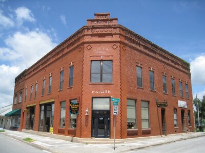A historic red brick building with large windows stands on a street corner under a blue sky with clouds in Lafayette County, Florida. Signs and awnings adorn the structure, and a street sign is visible at the intersection. The building boasts decorative brickwork along the roofline.