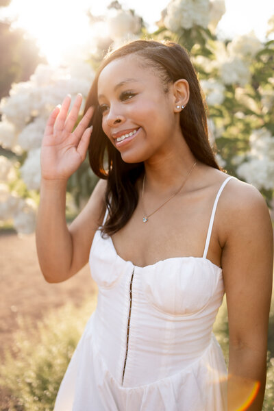 a teenage girl tucks her hair behind her ear as she stands in front of  a blooming white hydrangea tree with soft sunlight pouring into the image