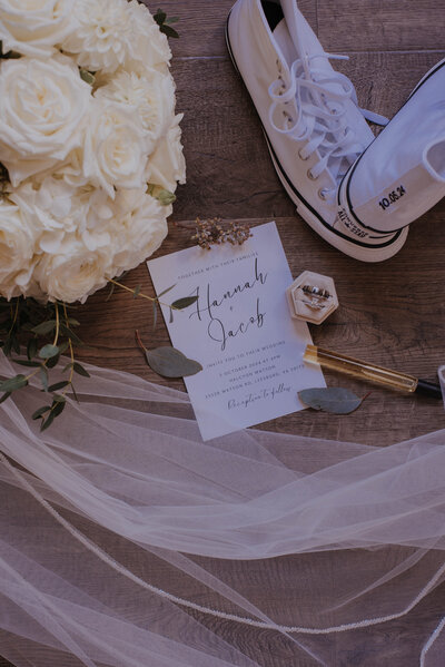 A wedding invitation on a wooden floor with white sneakers, a bouquet of white roses, a veil, and a ring box captures the essence of a rustic celebration. Delicate jewelry and a pen are nearby, perfect for planning at event space rentals in Loudoun County.