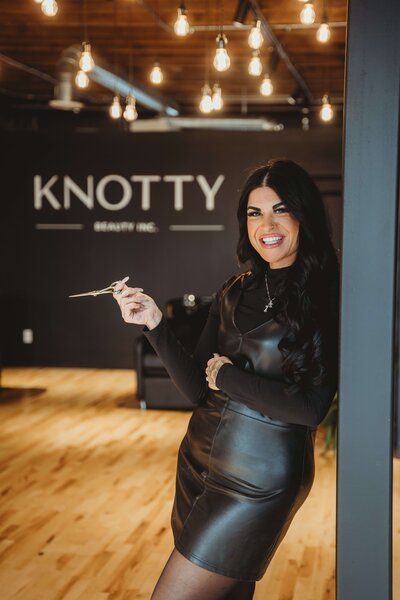 Santina leaning on a ledge wearing all black, smiling as she holds hair cutting scissors.