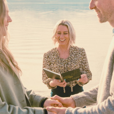 Celebrant Kirsty lakeside with the couple blurred in the foreground, holding hands. All are smiling.