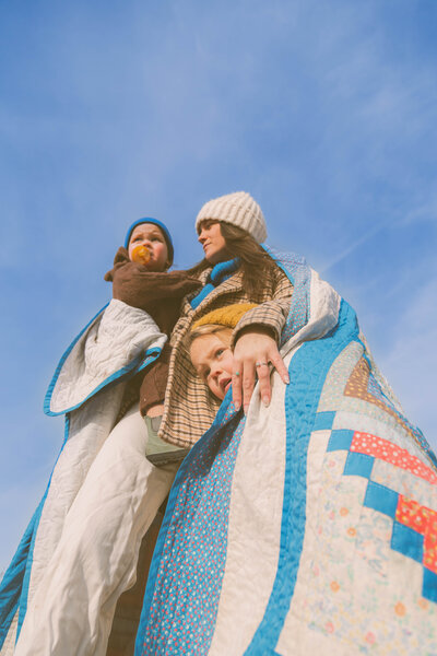 mom holds young son as daughter hugs her leg with a blue sky as the backdrop