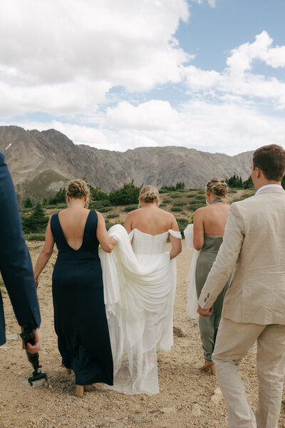 A bride, groom, and their family walking towards their ceremony spot in Colorado as the brides mom holds her dress up.