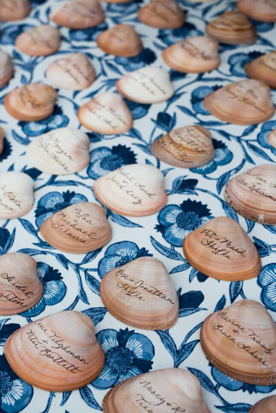 Clam shell escort cards with brown calligraphy on blue and white floral tablecloth for coastal wedding in Quogue, New York.