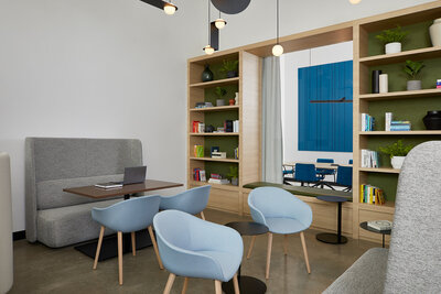 A bright, airy work area with numerous chairs, benches, and tables, and a large wood shelf  containing books, plants, and decorative art pieces