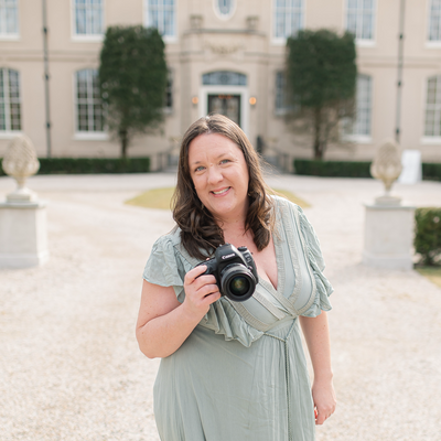 A woman with long brown hair holds a camera and stands in front of an elegant building with a courtyard. She is wearing a light green dress and is smiling warmly at the camera.