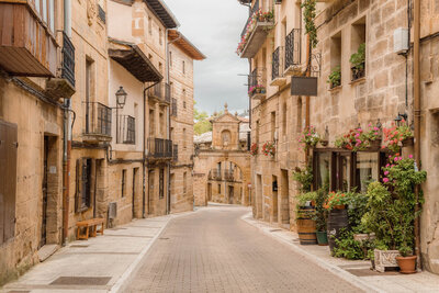 A charming, narrow cobblestone street in a historic European village, lined with stone buildings adorned with balconies and colorful flowers. A decorative archway is visible at the end of the street under a cloudy sky.