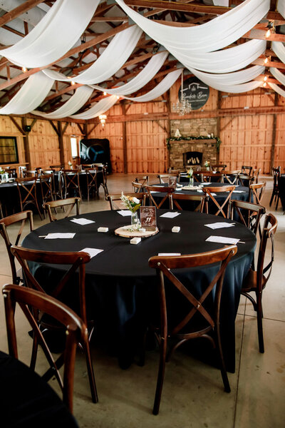 Round reception table with black tablecloth, wooden chairs, and small floral centerpiece in a rustic barn setting
