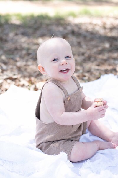 Baby in a brown romper sits on a white blanket outdoors, smiling in the sunlight. A joyful baby session by Moments in Light Photography.