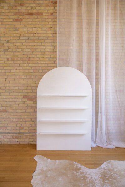 A six foot white arch with shelves set up against a brick wall.