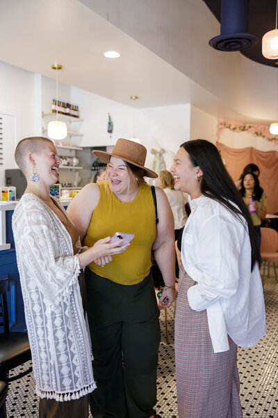Three women stand together in conversation and laughing at a networking event in Seattle. One woman on the left has a shaved head and wears a white lace cardigan. The woman in the middle sports a wide-brimmed hat and a yellow top, while the woman on the right has long dark hair and wears a white shirt.