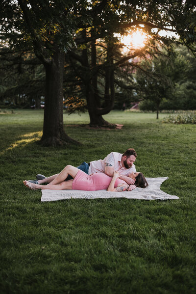 Pregnant wife in pink dress laying with husband in pink and blue on white blanket under trees in Baltimore Maryland.