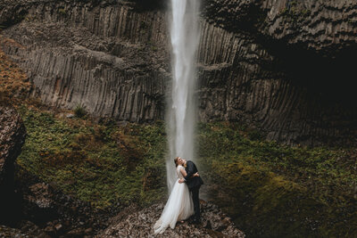 Bride and groom holding each other under a waterfall stealing a kiss from each other surround by the rocky landscape