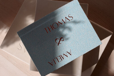 Blue wedding album with the names Thomas & Amelia debossed in copper on top of two other rose colored nubuck leather - Romero Album Design