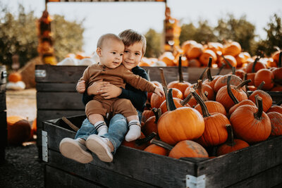 A playful autumn portrait of two siblings sitting among pumpkins at Curtis Orchard in Champaign, IL. Light By Zoey Photography offers seasonal family photography at Curtis Orchard and other locations in Champaign, Mahomet, and Urbana, perfect for capturing festive moments during the fall season.