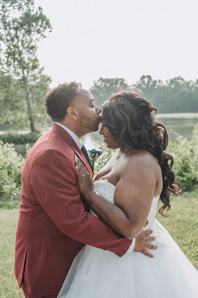 A groom in a red suit kisses the forehead of his bride, who is wearing a strapless white wedding dress, as they embrace outdoors at Bull Run Golf Club in Haymarket, Virginia. The background features lush greenery and a serene lake. Shot by Quinn Photography LLC.