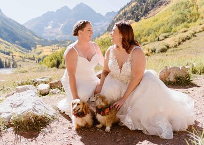 Two brides in wedding dresses smiling with their flower-collar dachshunds at their September elopement at Maroon Bells Amphitheater in Aspen, Colorado with golden fall foliage and mountains in the background