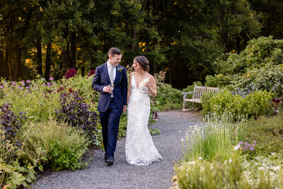 Bride and groom walk down a gravel path in a Virginia park with drinks in hand
