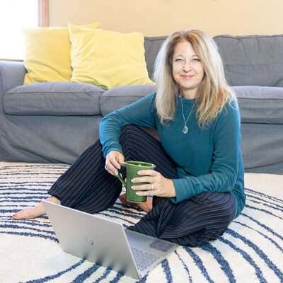 Cyndi Stewart sits barefoot on a rug with a laptop and green mug, smiling warmly in a cozy, casual home setting.