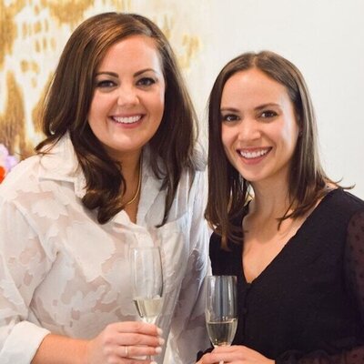 Two women are standing close together, each holding a glass of champagne. One wears a white blouse and the other wears a black top. Both are smiling and appear to be in a well-lit, elegant setting.