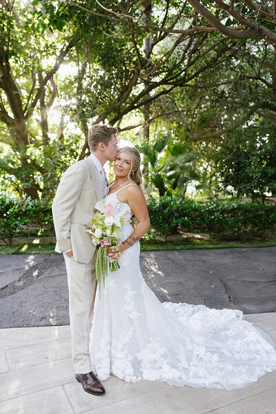 Bride and groom standing groom kissing her on her temple and bride holding her bouquet 