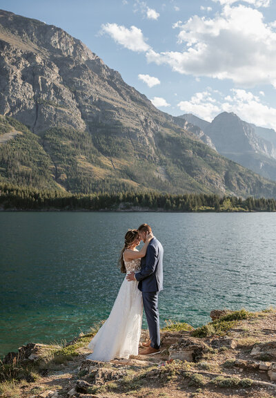A couple shares an intimate embrace beside a sparkling alpine lake with towering mountain peaks in the background, bathed in golden afternoon light during their Glacier National Park elopement, captured by Sydney Breann Photography.