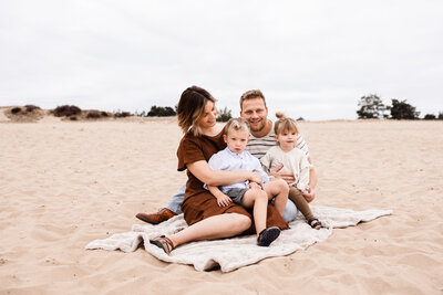 Gezinsshoot op een zandvlakte bij eendenmeer bergen papa mama zoon en dochter zitten op kleedje beige bruine kleren