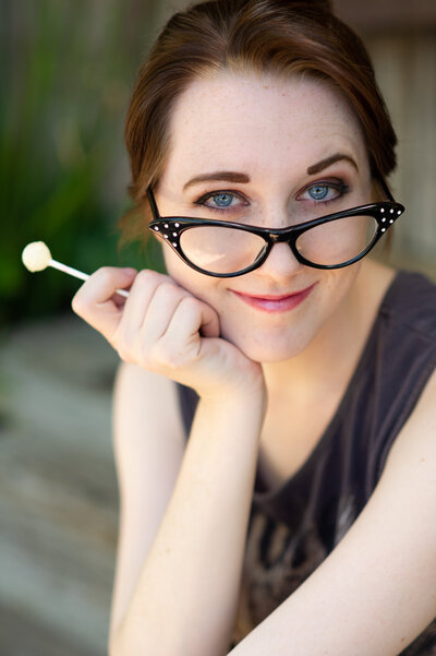A teenage actress with brown hair and piercing blue eyes, wearing quirky glasses and holding a lollipop for a fun headshot photo for commercial work.
