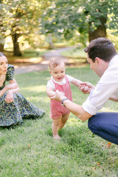 A toddler smiles while his father helps him to walk during a Rochester NY newborn photography and family session.