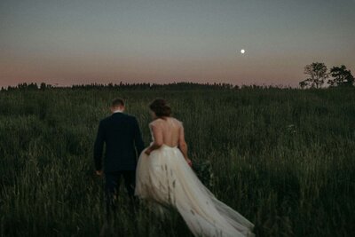 bride and groom walk together during blue hour featuring the moon and a field of greenery
